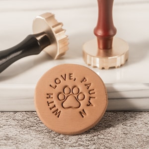 May include: A collection of three brass and wood wax seal stamps. One stamp has a paw print design with the words "WITH LOVE, PAULA N" around it. The other two stamps have different decorative designs.