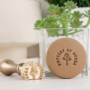 May include: A brass-colored wax seal stamp with a wooden handle and a floral design. A round, tan-colored wax seal with the text "POTTERY BY BREZA" surrounds a floral illustration. A small succulent plant in a glass container is in the background.