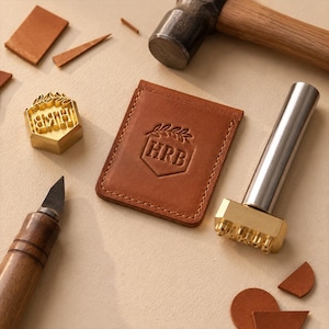 Puede incluir: Un tarjetero de cuero color canela con las letras HRB grabadas dentro de un diseño de escudo y hojas, acompañado de un sello de latón con el mismo logotipo, un mazo de metal, un cúter y pequeños trozos de cuero sobre una superficie clara.