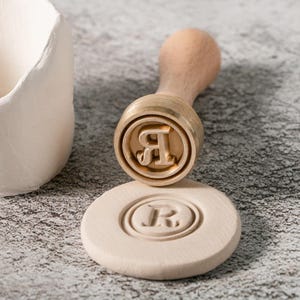 May include: A wooden handled brass stamp with the letter "R" in a circular design. The stamp is used to create an impression in clay.