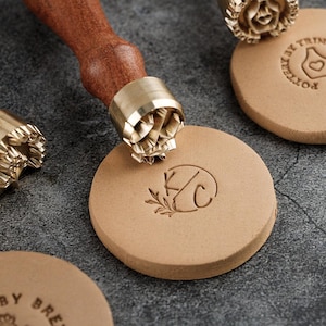 May include: A close-up of leather stamps with wooden handles and brass stamp heads. One stamp is pressed into a round piece of leather, creating a "KC" monogram with a leaf design. Other stamps and leather pieces are visible.