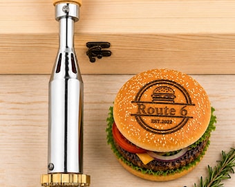 May include: A silver metal branding iron with a light wood handle positioned next to a hamburger bun branded with the text Route 6 EST.2022. The burger features a patty, lettuce, tomato, cheese, and onion, with a sprig of rosemary and peppercorns.