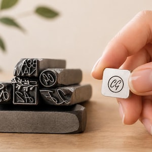 May include: A collection of silver-toned metal stamping tools with various engraved designs, including a square stamp featuring the letters CG inside a circle, a rectangular stamp with a floral motif and the letters AZ, and other letter stamps.