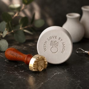 May include: A brass pottery stamp with a dark wood handle featuring the text Pottery By Jason, positioned next to a circular white clay disc embossed with a vase of flowers and the text WITH LOVE BY JASON in a circular arrangement.
