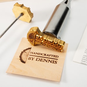 Puede incluir: Una herramienta de pirograbado con una cabeza de latón que dice "HANDCRAFTED BY DENNIS". La herramienta se utiliza para crear diseños personalizados de madera quemada.