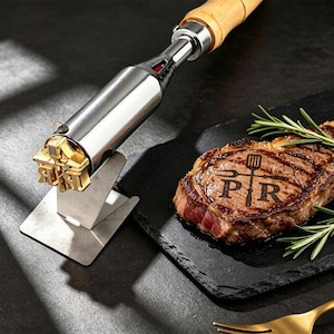 May include: A metal branding iron with a wooden handle and a gold-toned head featuring the letters P and R with a spatula and fork icon. The iron rests on a metal stand next to a cooked steak branded with the same P R spatula and fork design.