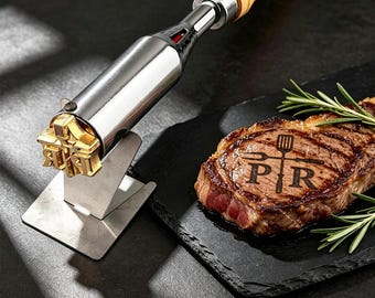 May include: A metal branding iron with a wooden handle and a gold-toned head featuring the letters P and R with a spatula and fork icon. The iron rests on a metal stand next to a cooked steak branded with the same P R spatula and fork design.