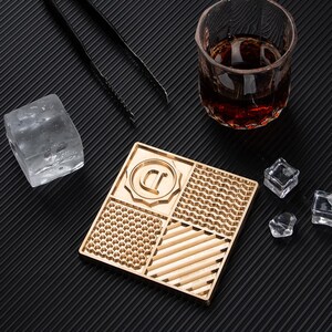 May include: A gold-colored square coaster with four different textured patterns. The coaster has a raised letter 'D' in a circle in the center. The coaster is on a black surface with ice cubes and a glass of brown liquid.