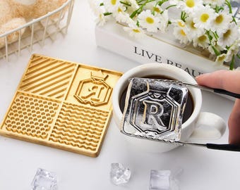 May include: A gold-coloured wooden stamp with four patterned sections and a logo. A large ice cube with the letter "R" is held with tongs over a white mug of coffee. Several ice cubes are scattered around.