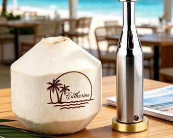 May include: A white coconut with a brown laser-engraved logo featuring two palm trees, waves, and the name Catherine, positioned next to a tall, cylindrical stainless steel coconut opener tool with a wooden handle and a brass-colored base.