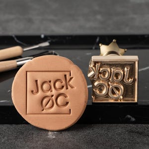 May include: A round, light brown clay stamp with the text "Jack ØC" inside a square. A gold-colored metal stamp with the same text and a crown handle is next to the clay stamp. Crafting tools are in the background.
