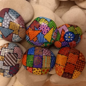 Könnte beinhalten: Sechs handbemalte Steine mit farbenfrohen Patchwork-Designs. Jeder Stein weist ein einzigartiges Muster aus Quadraten und Formen in verschiedenen Farben auf, darunter Blau, Orange, Grün und Rot. Die Steine werden auf einem Bett aus glatten, hellen Steinen präsentiert.
