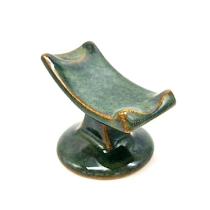 Puede incluir: Un soporte de cerámica verde con una parte superior curva y una base redonda. El soporte tiene un esmalte moteado y un borde dorado.