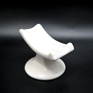 May include: A white ceramic stand with a curved top and a round base. The stand is designed to hold a small object, such as a phone or a tablet.