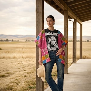 May include: A black t-shirt featuring a white graphic of Dolly Parton and the text DOLLY, worn under a pink, blue, and green patchwork kimono. The outfit includes dark blue denim jeans and a light beige wide-brimmed hat held in one hand.