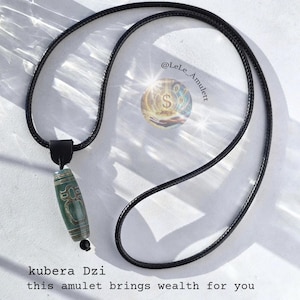 Può includere: Collana con ciondolo amuleto Kubera Dzi e cordino nero. Il ciondolo cilindrico presenta un design verde e bianco. Il testo "kubera Dzi" e "this amulet brings wealth for you" è in basso nell'immagine.