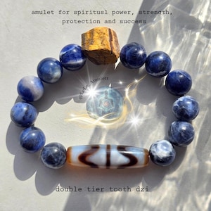 Peut inclure: Un bracelet de perles composé de sodalite bleue et blanche, d'une pierre œil-de-tigre brute et d'une perle dzi cylindrique blanche et brune à double dent. Le texte indique : amulet for spiritusl power, strength, protection and success.