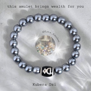 Könnte beinhalten: Ein silbernes Perlenarmband mit einem schwarzen und weißen Dzi-Perlenanhänger. Der Anhänger hat ein Symbol darauf und der Text "Kubera Dzi" ist unter dem Armband geschrieben. Der Text "this amulet brings wealth for you" ist über dem Armband geschrieben. Der Text "@LeLe_Amulet" ist auf dem Armband geschrieben.