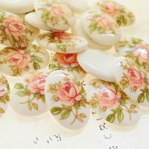 May include: A collection of oval-shaped, white ceramic buttons with a floral design. The buttons feature pink roses and green leaves on a white background.