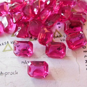 May include: A group of pink, faceted, rectangular glass gems. The gems are scattered on a white surface with black text.