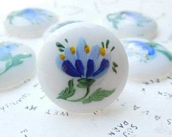 Puede incluir: Un conjunto de seis cabujones de vidrio blanco con flores pintadas a mano en azul y amarillo. Los cabujones son redondos y tienen un acabado liso y brillante.