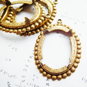 以下が含まれることがあります： アンティークゴールドカラーの金属製オーバルペンダントフレームのクローズアップ。フレームは、小さな丸い装飾と吊り下げ用のループを備えた、詳細でテクスチャ加工されたデザインです。フレームは、薄いテキストのある白い背景にあります。