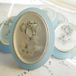 May include: Three light blue oval-shaped decorative trinkets with gold trim and a white floral design. Each trinket features a single rose with leaves.