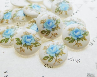 May include: A collection of white, round ceramic buttons. Each button features a blue rose with green leaves and gold accents. The buttons are scattered on a textured, off-white surface.