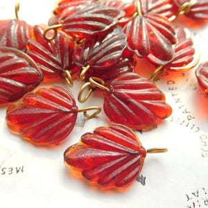 May include: A collection of red glass leaf-shaped beads with gold-colored wire loops. The beads have a ribbed texture and are scattered on a white surface with printed text.