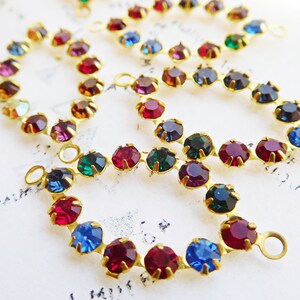 May include: A close-up of a gold chain with colorful rhinestones. The chain has two loops at the end. The rhinestones are red, blue, green, and purple.