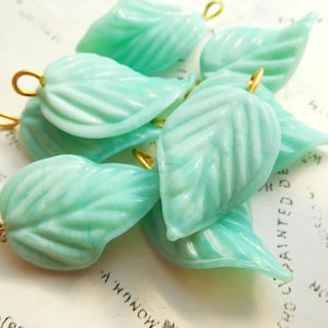 May include: A collection of light teal-colored leaf-shaped beads with gold-colored loops. The beads have a textured, veined design and are scattered on a light-colored surface.