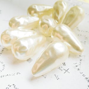 May include: A group of pearlescent white teardrop-shaped beads, some with a slight iridescent sheen, arranged on a white surface with black writing.