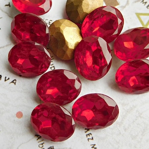 May include: A group of oval-shaped, red glass rhinestones. The rhinestones have a faceted design and are scattered on a white surface.