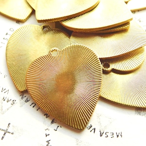 May include: A collection of heart-shaped brass pendants. Each pendant has a textured, sunburst design radiating from the center. The pendants have a small hole at the top for stringing. The background is a white surface with printed text.