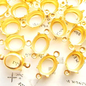 May include: Close-up of several gold-colored bezel settings for jewelry making. Each setting has an oval-shaped cavity and small loops for attaching to other components. The settings are arranged on a patterned surface.