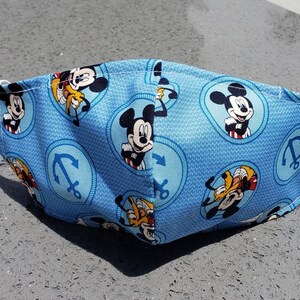 Op de afbeelding: Een blauw stoffen mondkapje met een herhalend patroon van Mickey Mouse en Donald Duck-figuren, ankers en een blauw-witte stippenachtergrond.