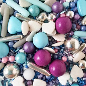 May include: A close-up of a mix of colourful sprinkles. The sprinkles include silver metallic balls, light blue rods, heart-shaped pieces, and various sizes of round sprinkles in shades of blue, purple, and white. The image has a bright, cheerful aesthetic.