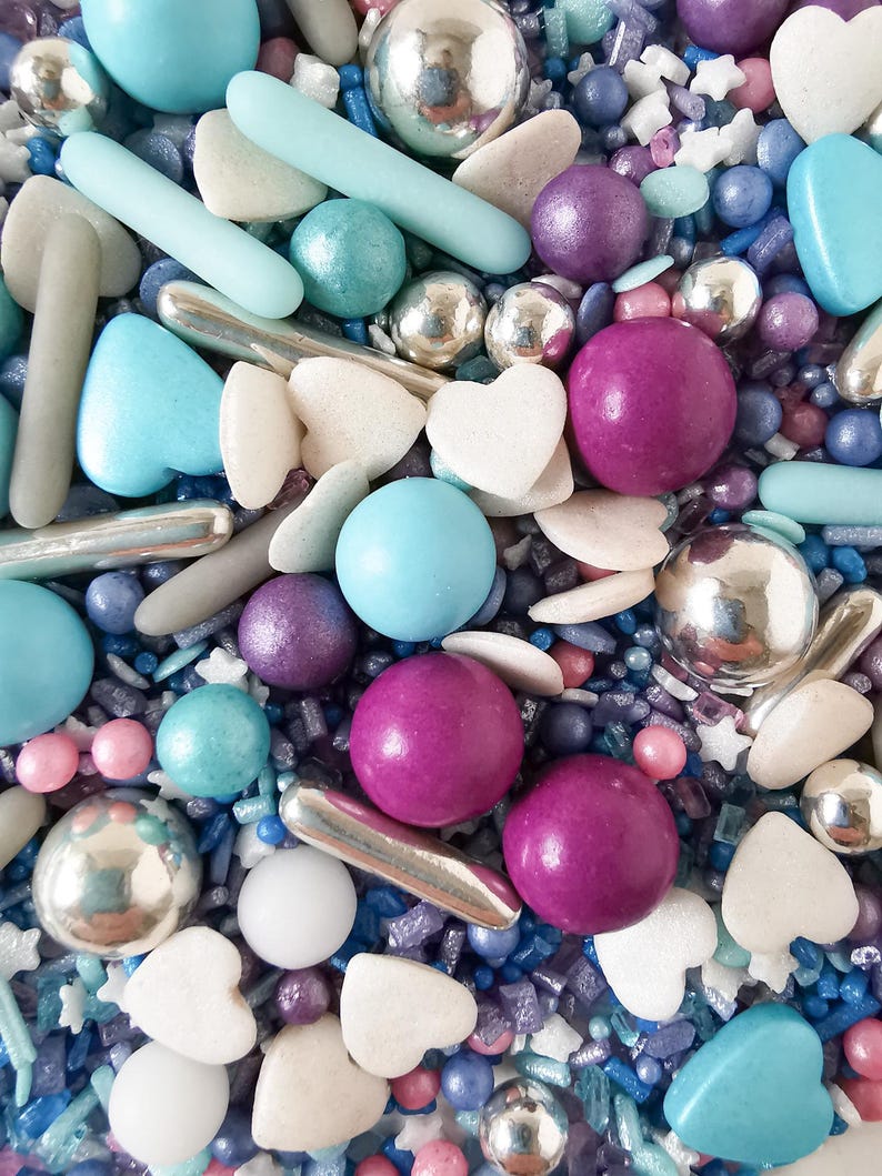May include: A close-up of a mix of colourful sprinkles. The sprinkles include silver metallic balls, light blue rods, heart-shaped pieces, and various sizes of round sprinkles in shades of blue, purple, and white. The image has a bright, cheerful aesthetic.