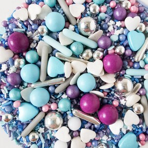 May include: A vibrant assortment of sprinkles, featuring blue, purple, and pink spheres, silver dragees, white hearts, and star-shaped sprinkles. The sprinkles are arranged in a circular design, creating a festive and decorative appearance.