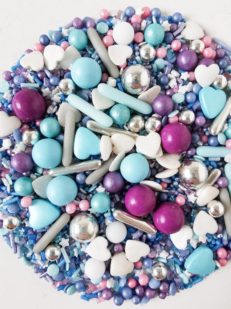 May include: A vibrant assortment of sprinkles, featuring blue, purple, and pink spheres, silver dragees, white hearts, and star-shaped sprinkles. The sprinkles are arranged in a circular design, creating a festive and decorative appearance.