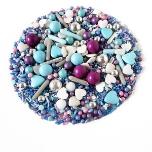 May include: A circular arrangement of colourful sprinkles and edible decorations. The mix includes blue, purple, silver, and white elements, such as round sprinkles, heart shapes, and small spheres. The sprinkles are on a white background.