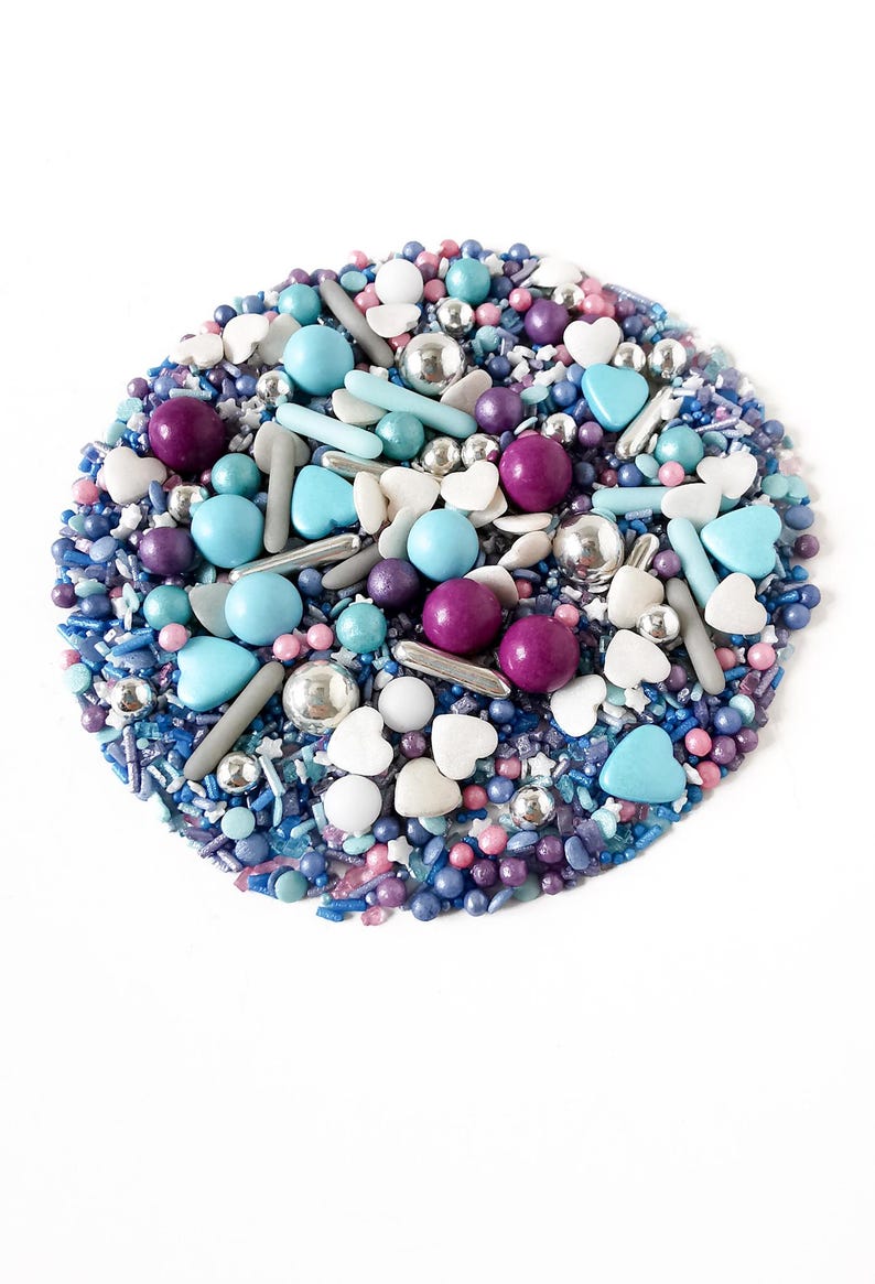 May include: A circular arrangement of colourful sprinkles and edible decorations. The mix includes blue, purple, silver, and white elements, such as round sprinkles, heart shapes, and small spheres. The sprinkles are on a white background.