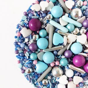 May include: A vibrant mix of sprinkles in blues, purples, pinks, and silver. The selection includes round, heart-shaped, and stick-shaped sprinkles, alongside small silver balls. The sprinkles are arranged on a white surface.