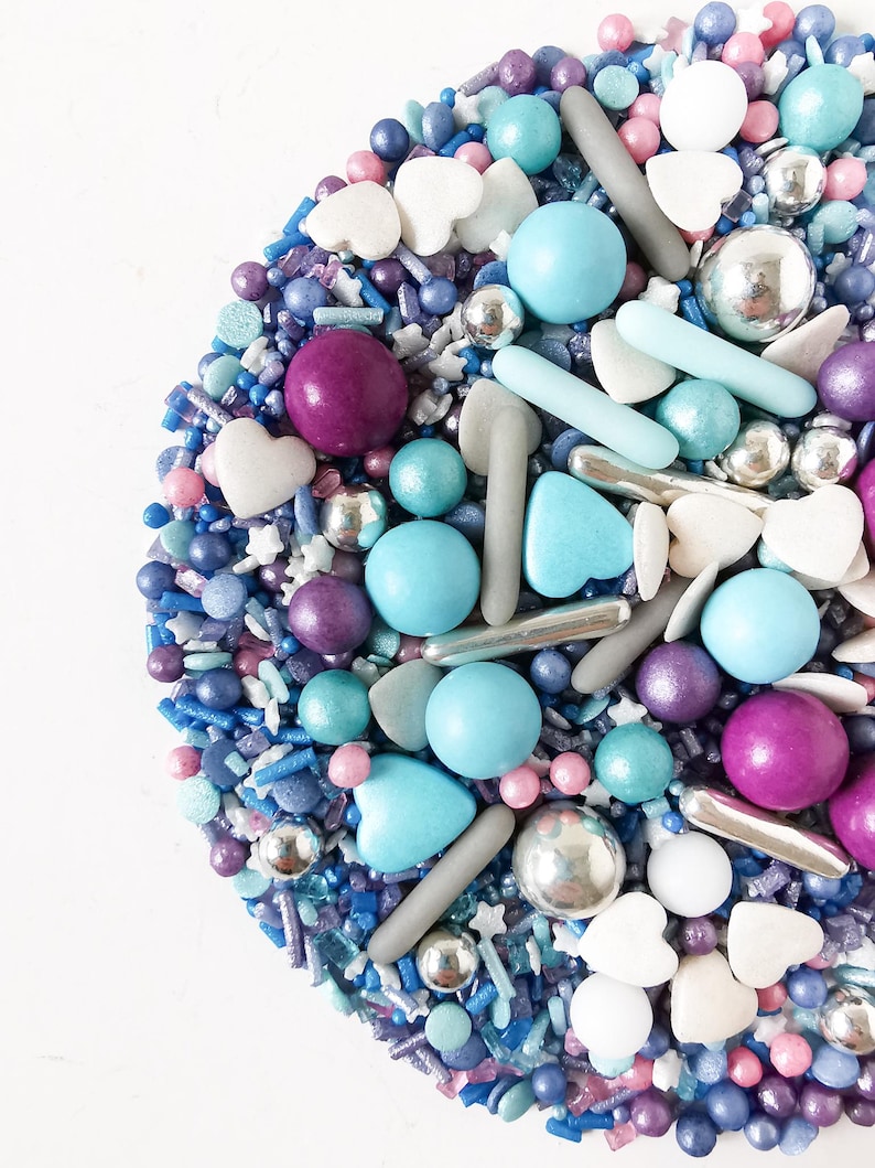 May include: A vibrant mix of sprinkles in blues, purples, pinks, and silver. The selection includes round, heart-shaped, and stick-shaped sprinkles, alongside small silver balls. The sprinkles are arranged on a white surface.