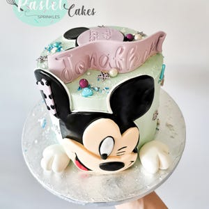 May include: A custom cake decorated with a Mickey Mouse design. The light green cake has black ears and a face, and a pink banner with the name "Jonah". It is decorated with sprinkles and edible pearls, and sits on a silver cake board.