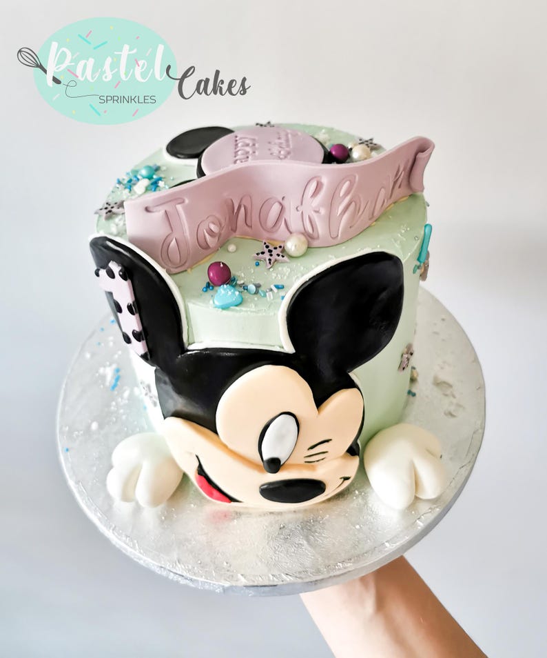 May include: A custom cake decorated with a Mickey Mouse design. The light green cake has black ears and a face, and a pink banner with the name "Jonah". It is decorated with sprinkles and edible pearls, and sits on a silver cake board.