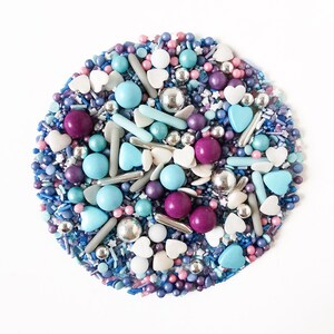 May include: A circular arrangement of colourful sprinkles, including blue, purple, pink, silver, and white. The sprinkles are a mix of shapes, including small balls, hearts, and long, thin pieces. The arrangement is on a white background.