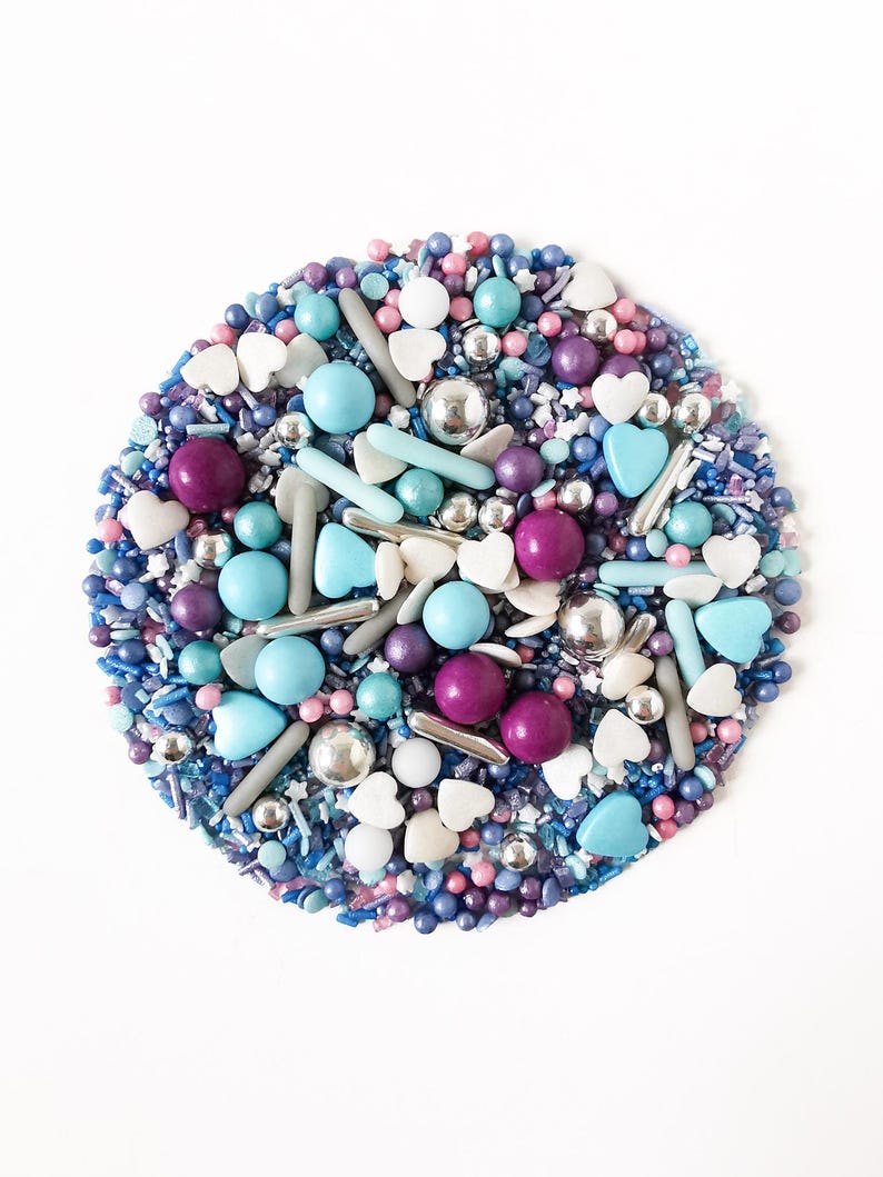 May include: A circular arrangement of colourful sprinkles, including blue, purple, pink, silver, and white. The sprinkles are a mix of shapes, including small balls, hearts, and long, thin pieces. The arrangement is on a white background.