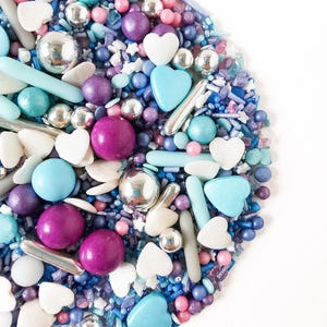 May include: A vibrant mix of sprinkles in blue, purple, pink, and silver. The selection includes round, heart, and rod-shaped sprinkles, ideal for decorating cakes and other treats. The sprinkles are displayed against a white backdrop.