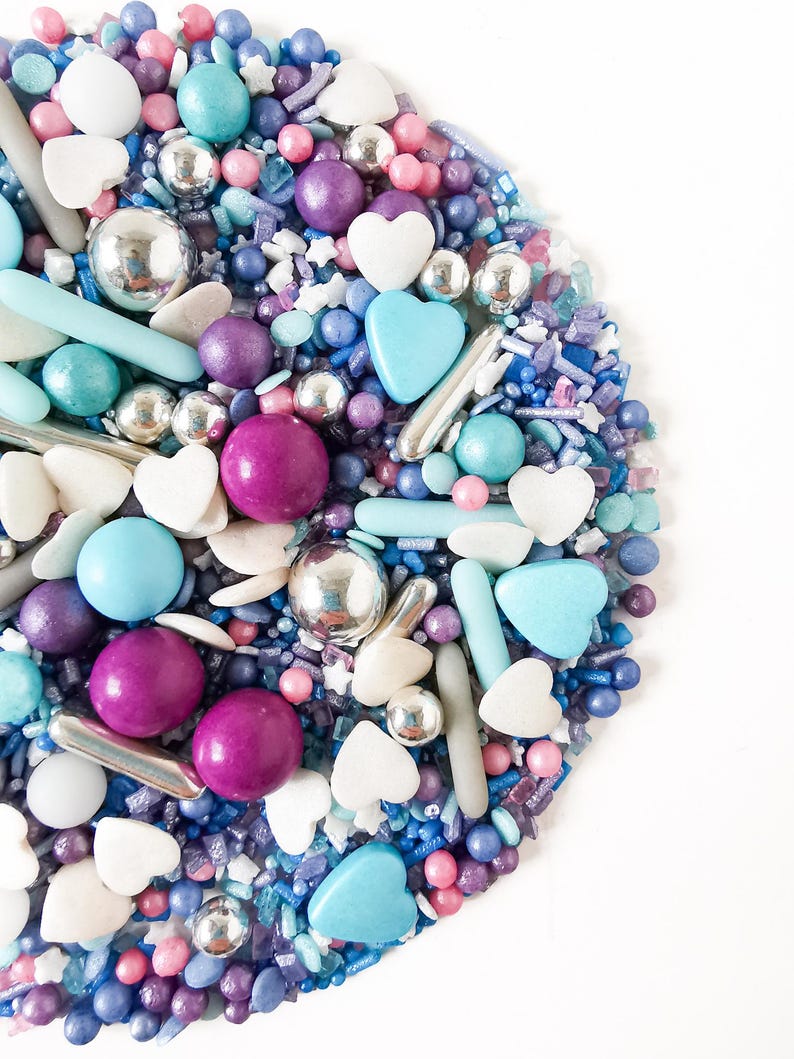 May include: A vibrant mix of sprinkles in blue, purple, pink, and silver. The selection includes round, heart, and rod-shaped sprinkles, ideal for decorating cakes and other treats. The sprinkles are displayed against a white backdrop.